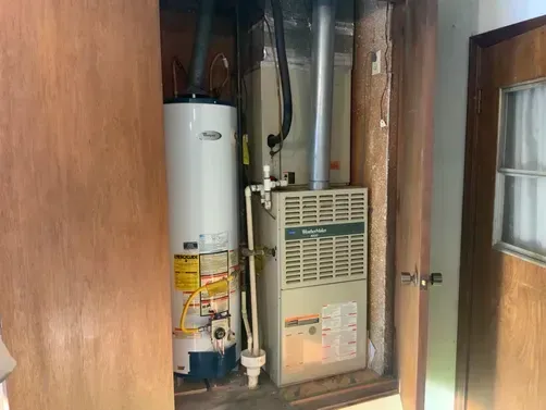 A white water heater and a beige furnace located inside a utility closet with wooden double doors.