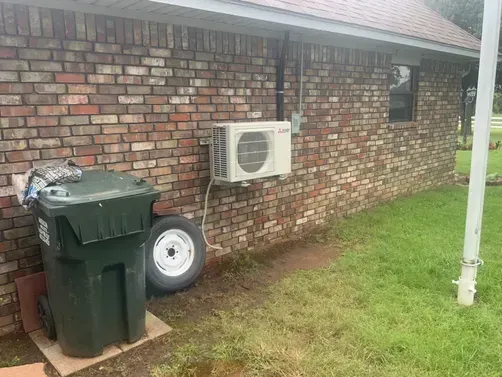 A brick exterior wall features an outdoor AC unit mounted above a green trash bin and a spare tire on a grass lawn.