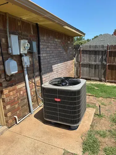A square Bryant HVAC condenser unit sits on a concrete pad next to a brick wall with exterior electrical components.