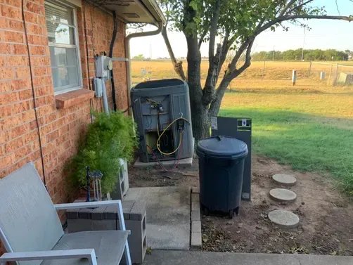 Outdoor scene with a brick house wall, an exposed HVAC unit, a grey trash can, and stepping stones on a grassy lawn.