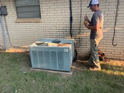 A technician in a baseball cap works on an outdoor HVAC unit against a brick wall.