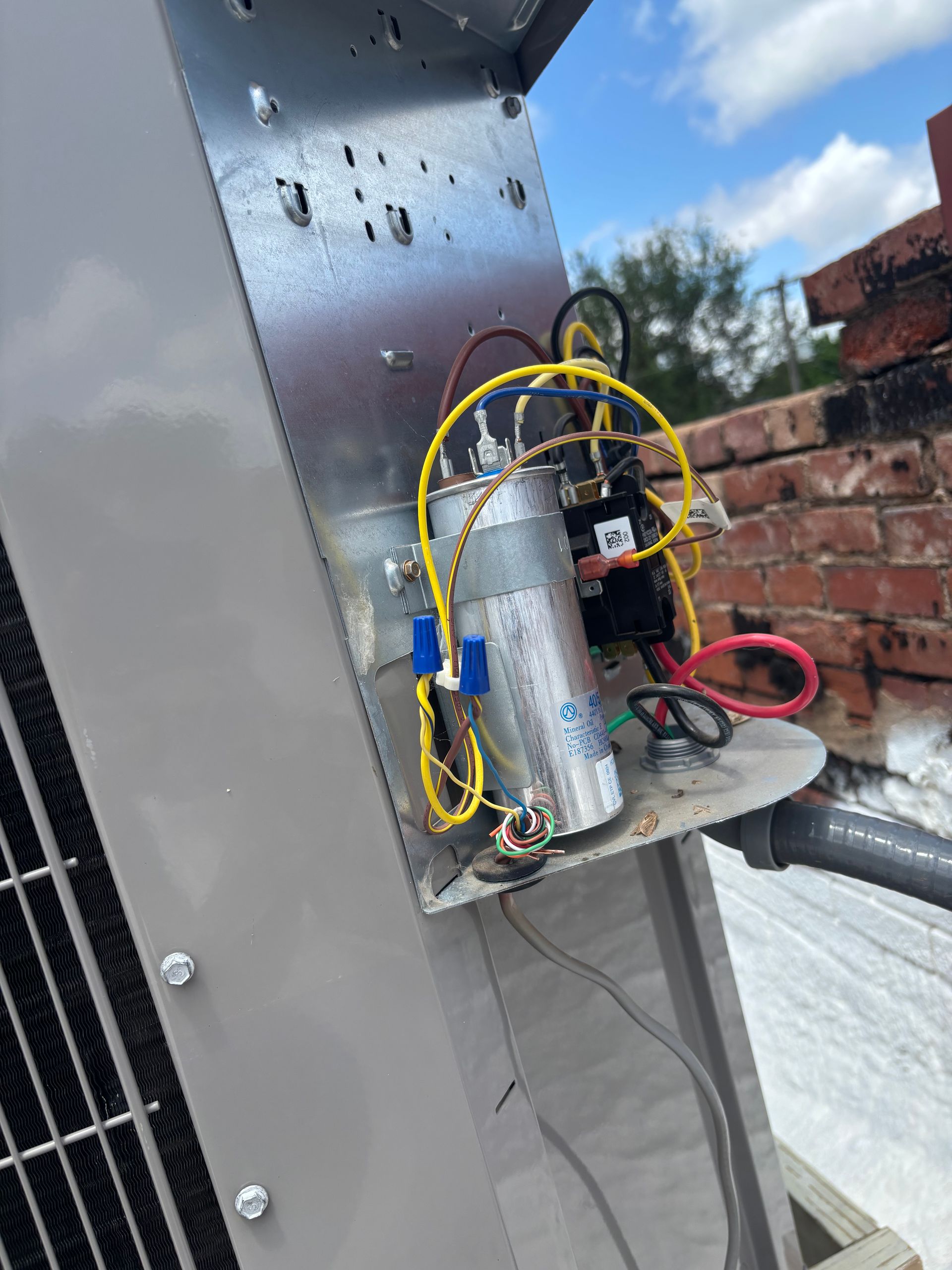 Open electrical service panel on an outdoor HVAC unit showing wiring and a capacitor against a brick chimney background.