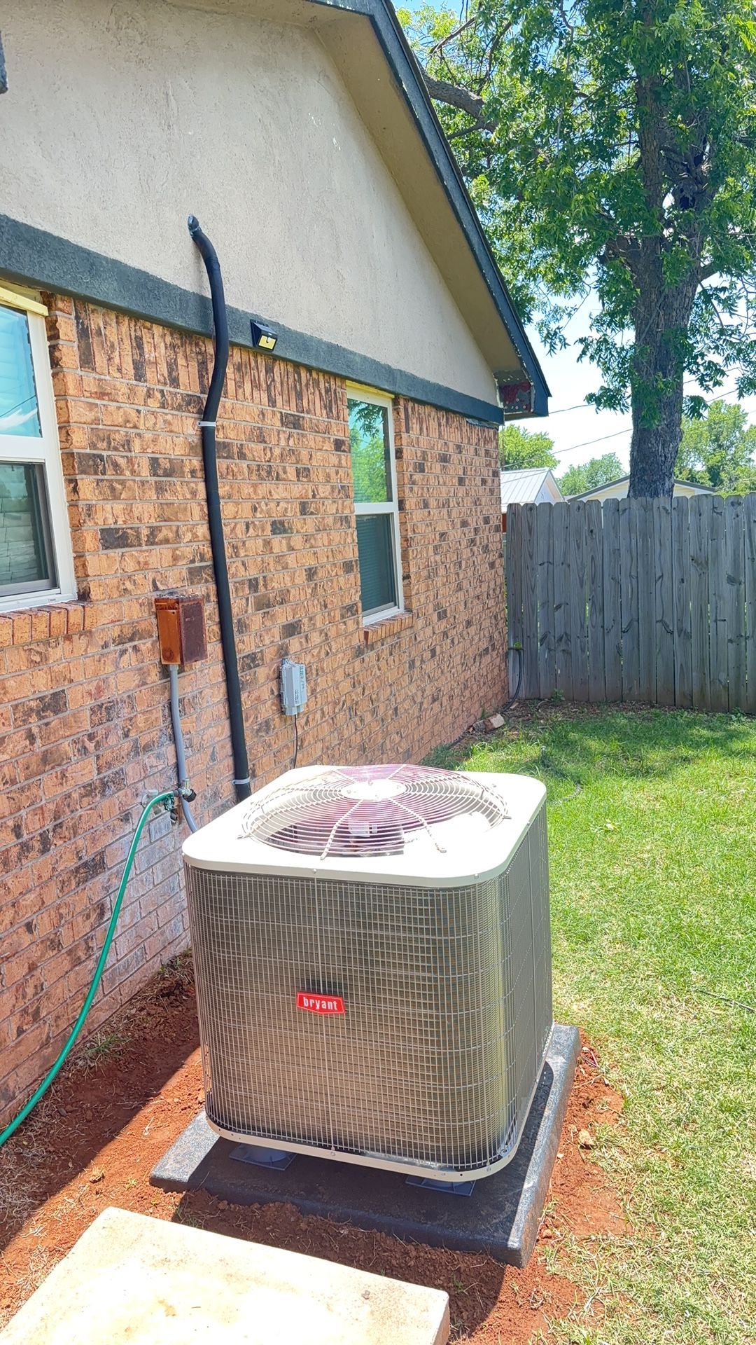 An HVAC condenser unit sits on a concrete pad outside a brick house, partially covered by a protective fabric mesh.