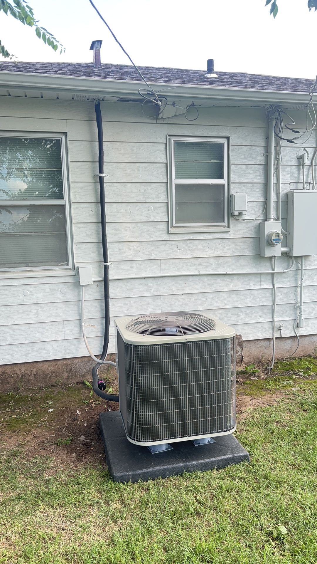 An outdoor central air conditioning unit sits on a concrete pad against the white-sided wall of a residential house.