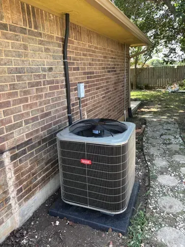 A Bryant brand air conditioning unit sits on a black pad against a brown brick house exterior next to a stone path.