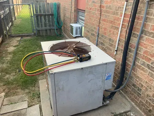 An HVAC outdoor condenser unit sits on a concrete pad next to a brick wall with refrigerant gauge hoses on top.