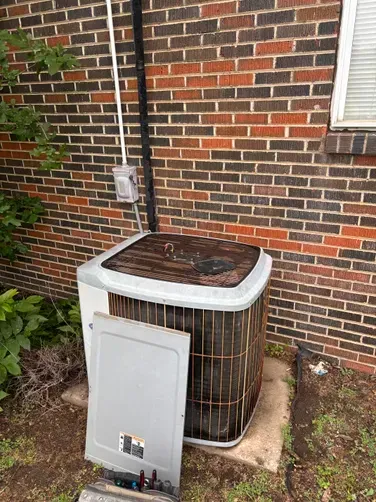 An outdoor HVAC condenser unit sits on a concrete pad against a brick wall with its electrical access panel removed.