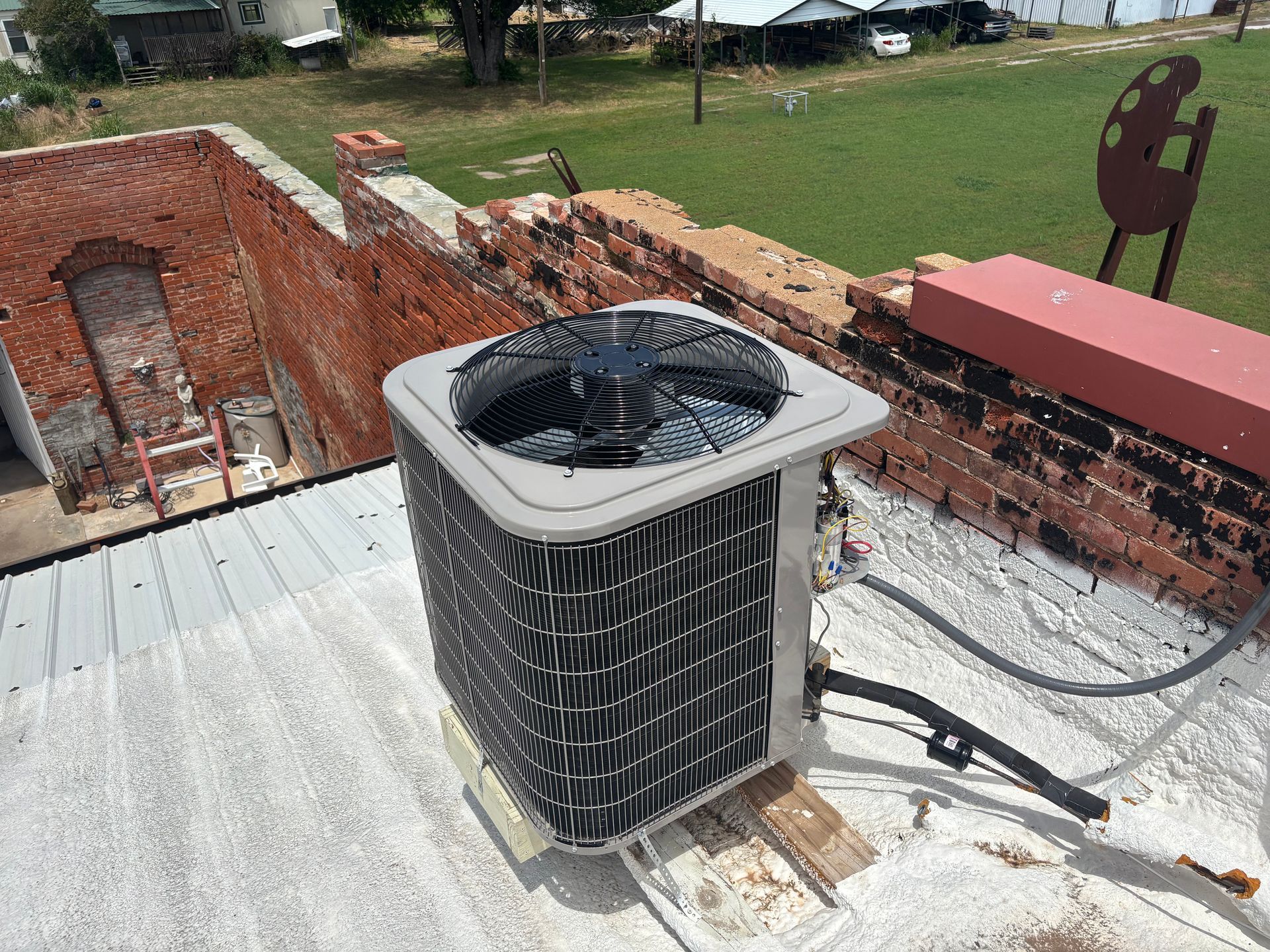 An air conditioning condenser unit sits on a flat, light-colored roof next to a weathered red brick wall.