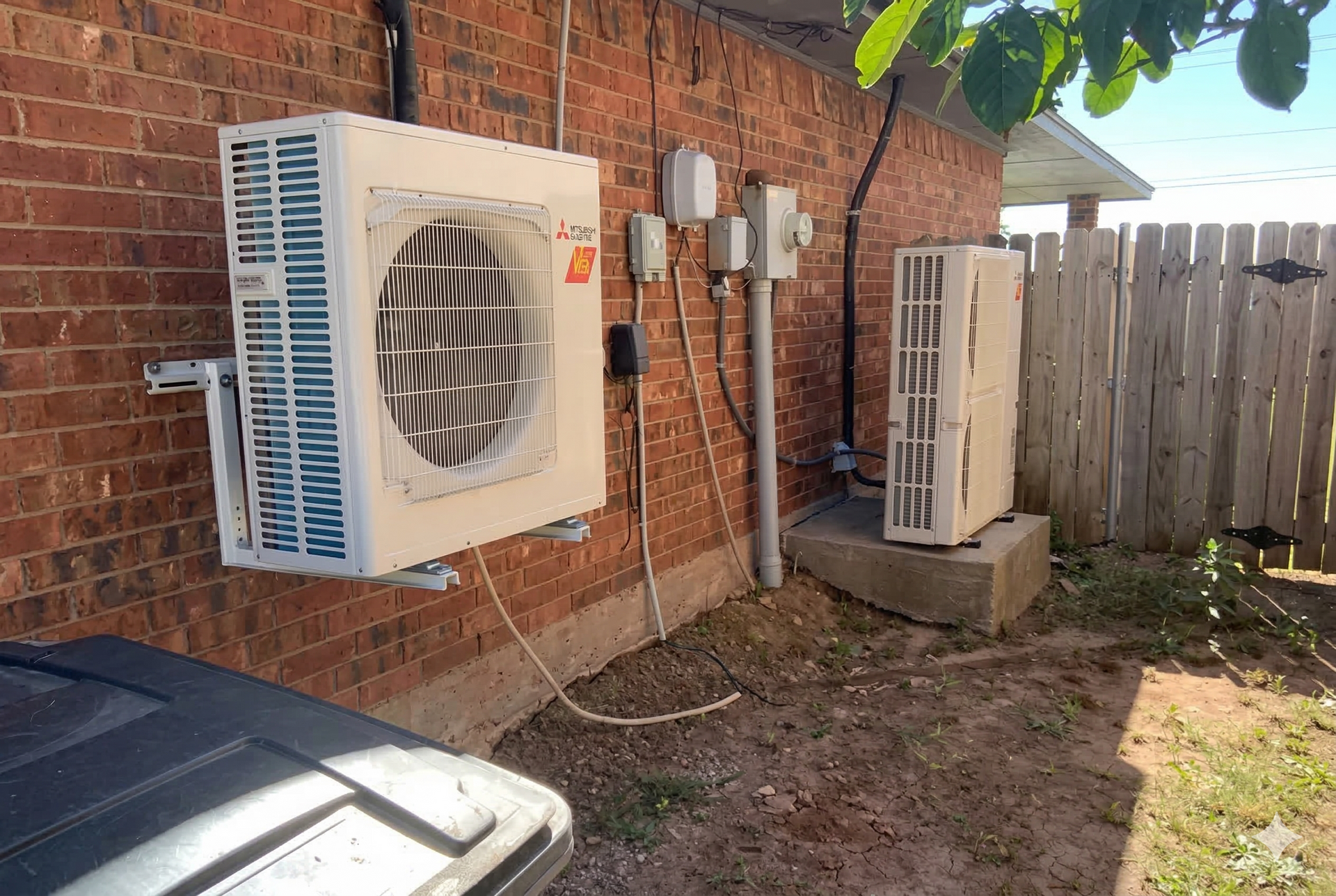 Two residential HVAC heat pump units installed on a brick wall exterior, with one wall-mounted and one on a concrete base.