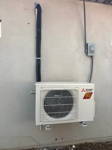 A beige Mitsubishi Electric outdoor air conditioning unit wall-mounted with black conduit and an electrical disconnect.