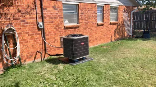 A Bryant air conditioning unit sits on a concrete pad next to a red brick house with three windows and a coiled hose.
