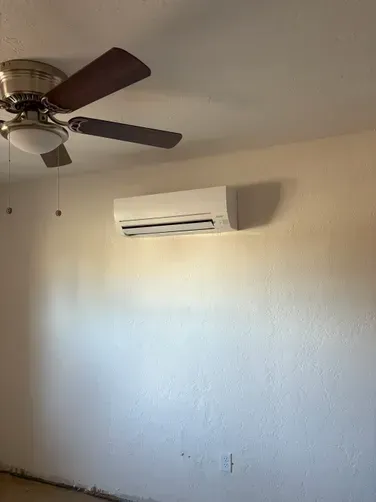 A white wall-mounted ductless air conditioning unit positioned below a ceiling fan with dark brown blades.
