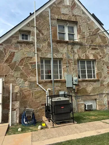 An outdoor HVAC unit mounted on a metal frame against a stone wall with utility pipes and electrical boxes nearby.