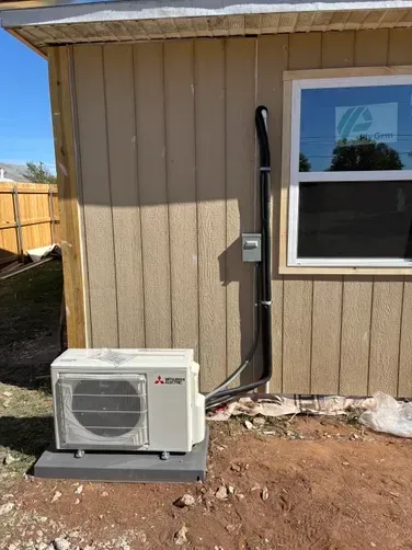 A beige mini-split heat pump unit sits on a concrete pad against a wooden shed wall, with black conduit running to a box.