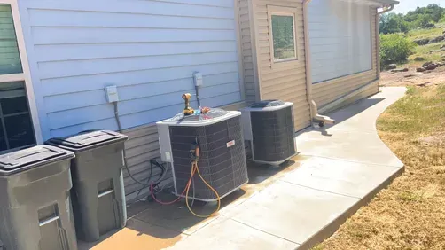 Two central air conditioning units sit on a concrete path outside a house with blue and tan siding.