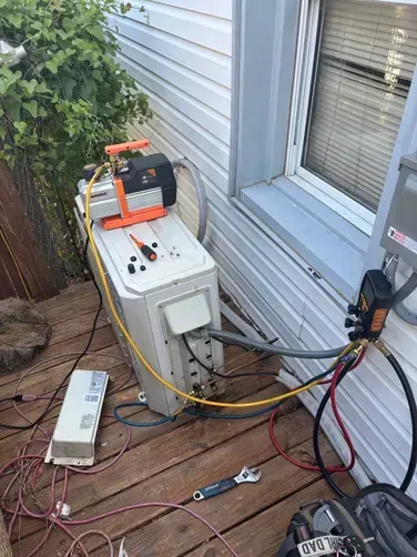 A vacuum pump is connected to an outdoor HVAC unit on a wooden deck to perform system maintenance.