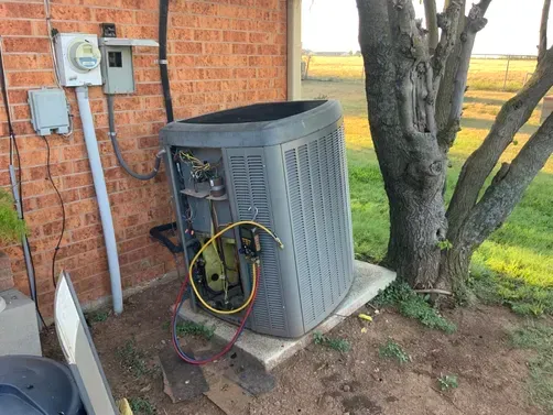 An outdoor HVAC unit sits on a concrete pad against a brick wall, with yellow and blue gauges attached to its side.