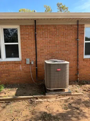 An outdoor Bryant air conditioning unit sits against a brick exterior wall near a window and two vertical pipes.