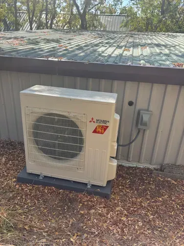 A beige Mitsubishi Electric outdoor HVAC unit sits on a dark base outside a building with ribbed metal siding.