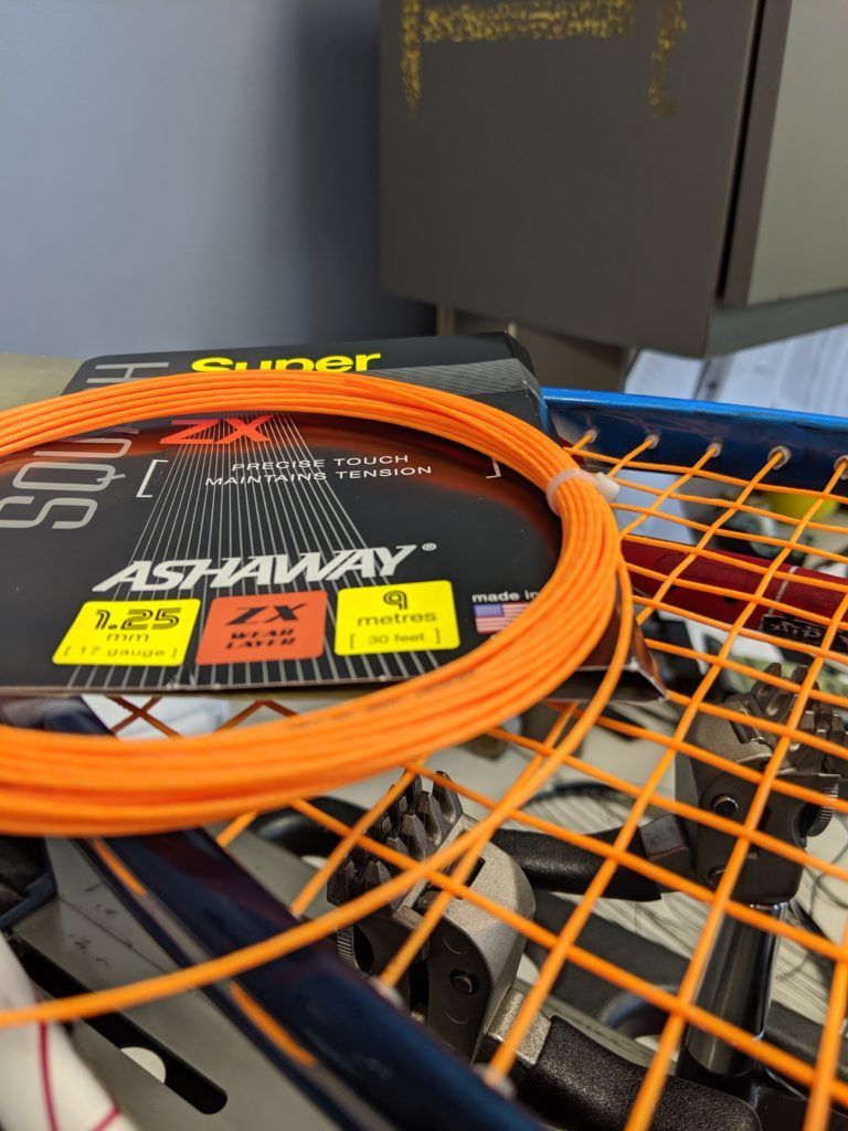An orange ashaway string is sitting on top of a tennis racket