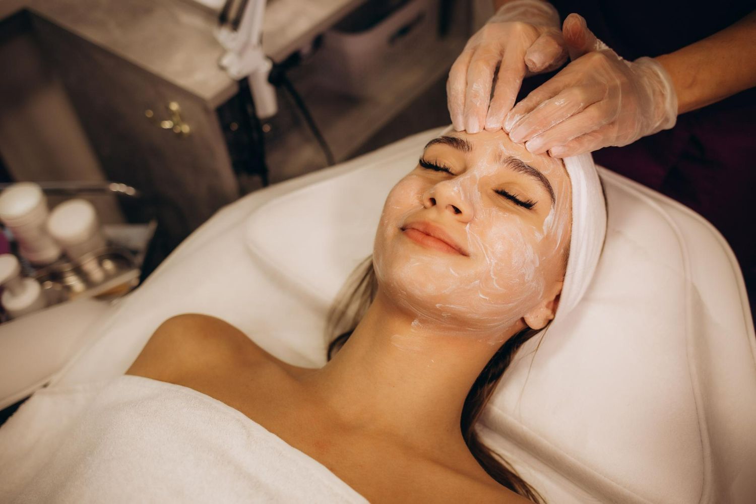 Woman receiving facial treatment at a spa; hands applying cream to face, eyes closed.