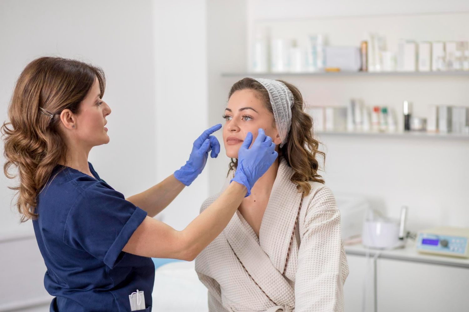A person in a robe has face examined by a person in blue gloves in a clinic room.