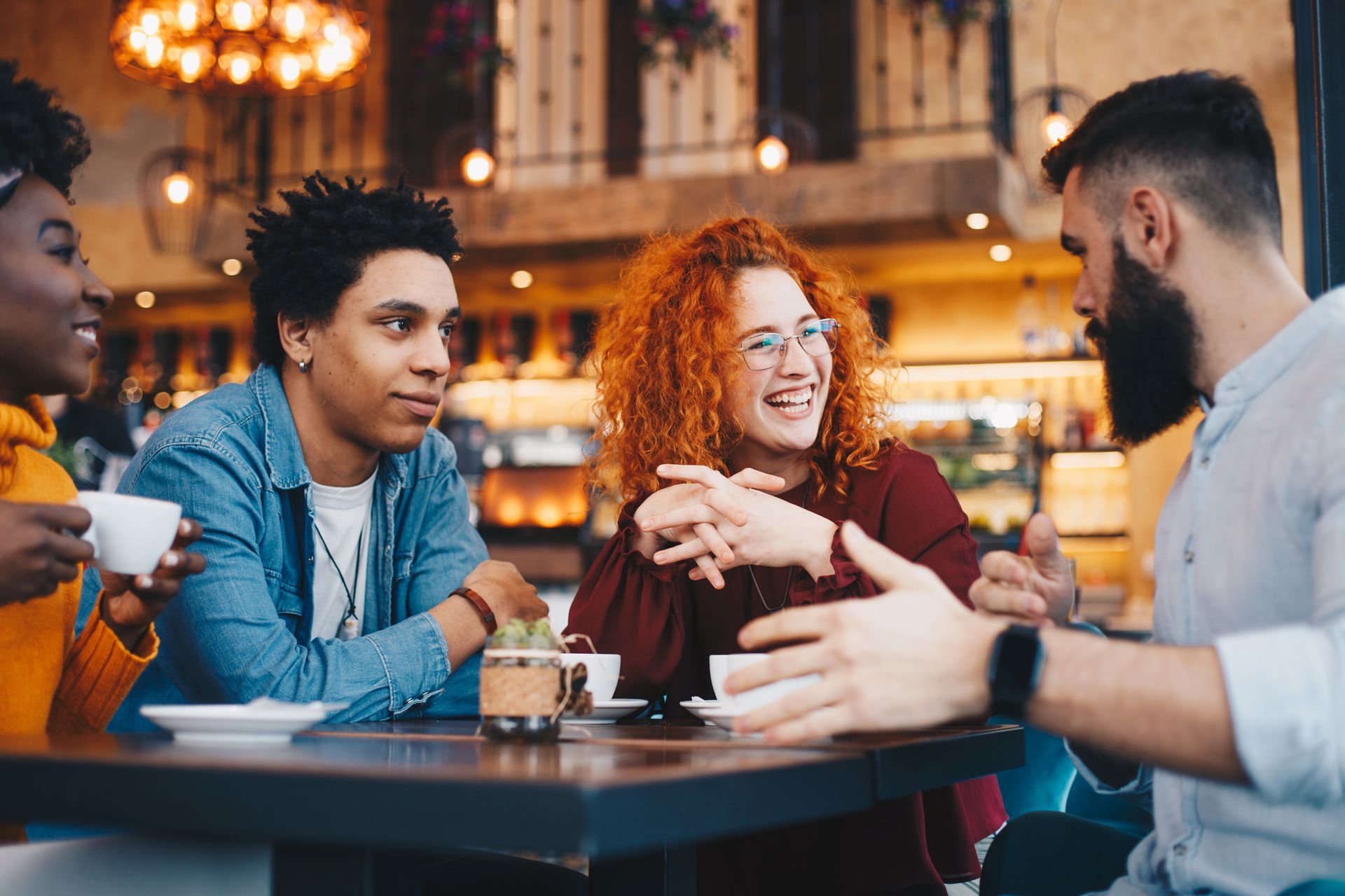 Friends laughing and talking around a table in a cafe, drinks in front of them.