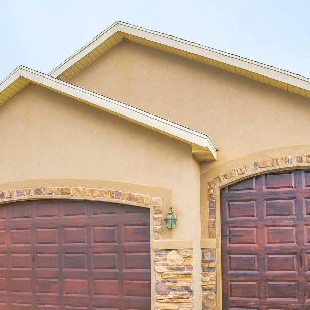 Two-car garage with brown wooden doors and stucco exterior. Light brown roof overhangs the doors.