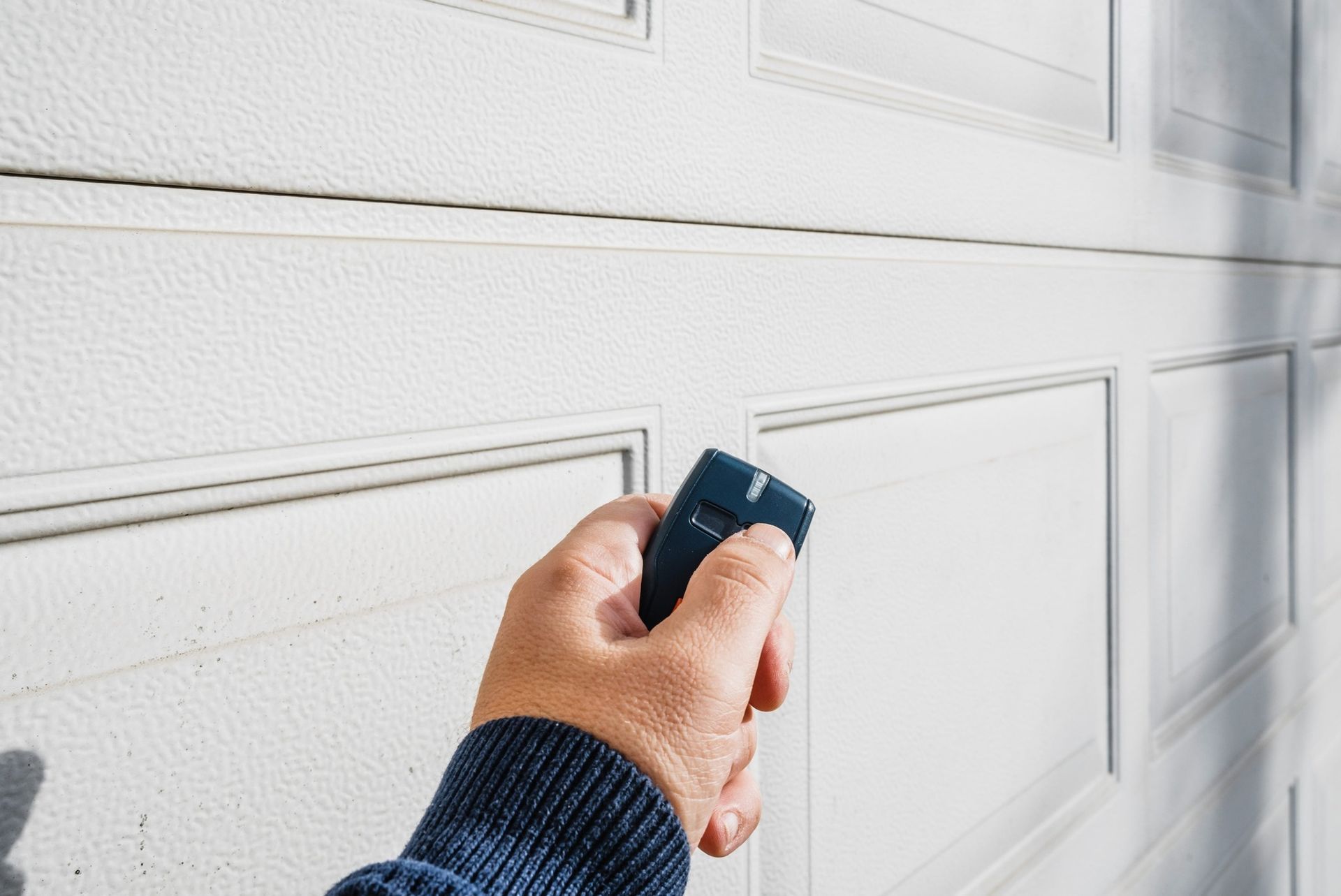Hand holding a garage door opener, aiming it at a white paneled garage door.