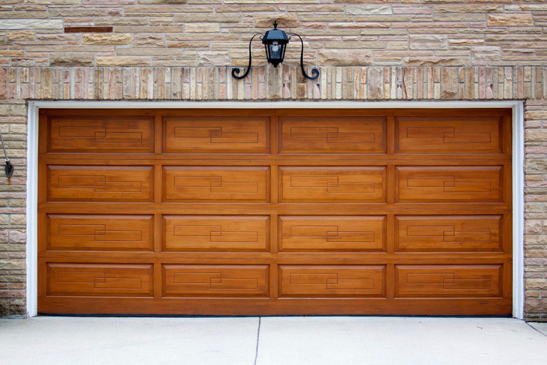 Wooden garage door with rectangular panels, set in a brick wall, with a decorative light fixture above.