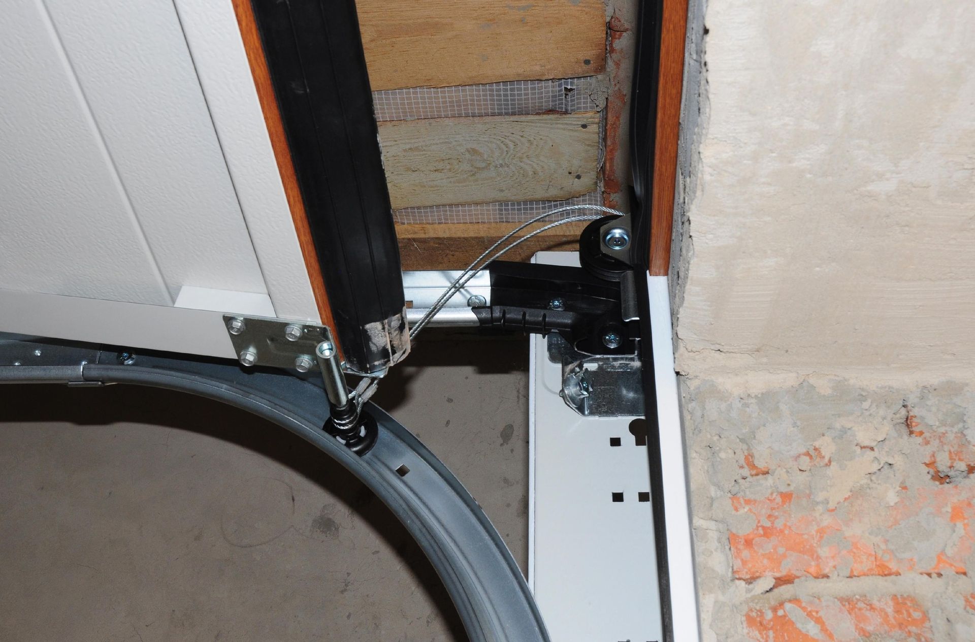 Garage door mechanism detail: curved track, cable, black rubber seal, brick wall, and mounting hardware.