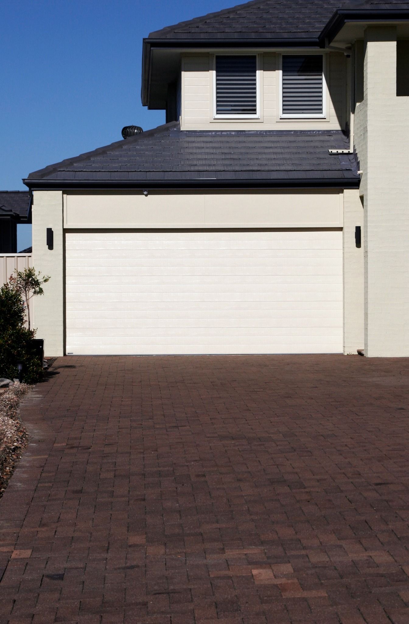 Beige house with a white garage door, dark brick driveway, and blue sky.