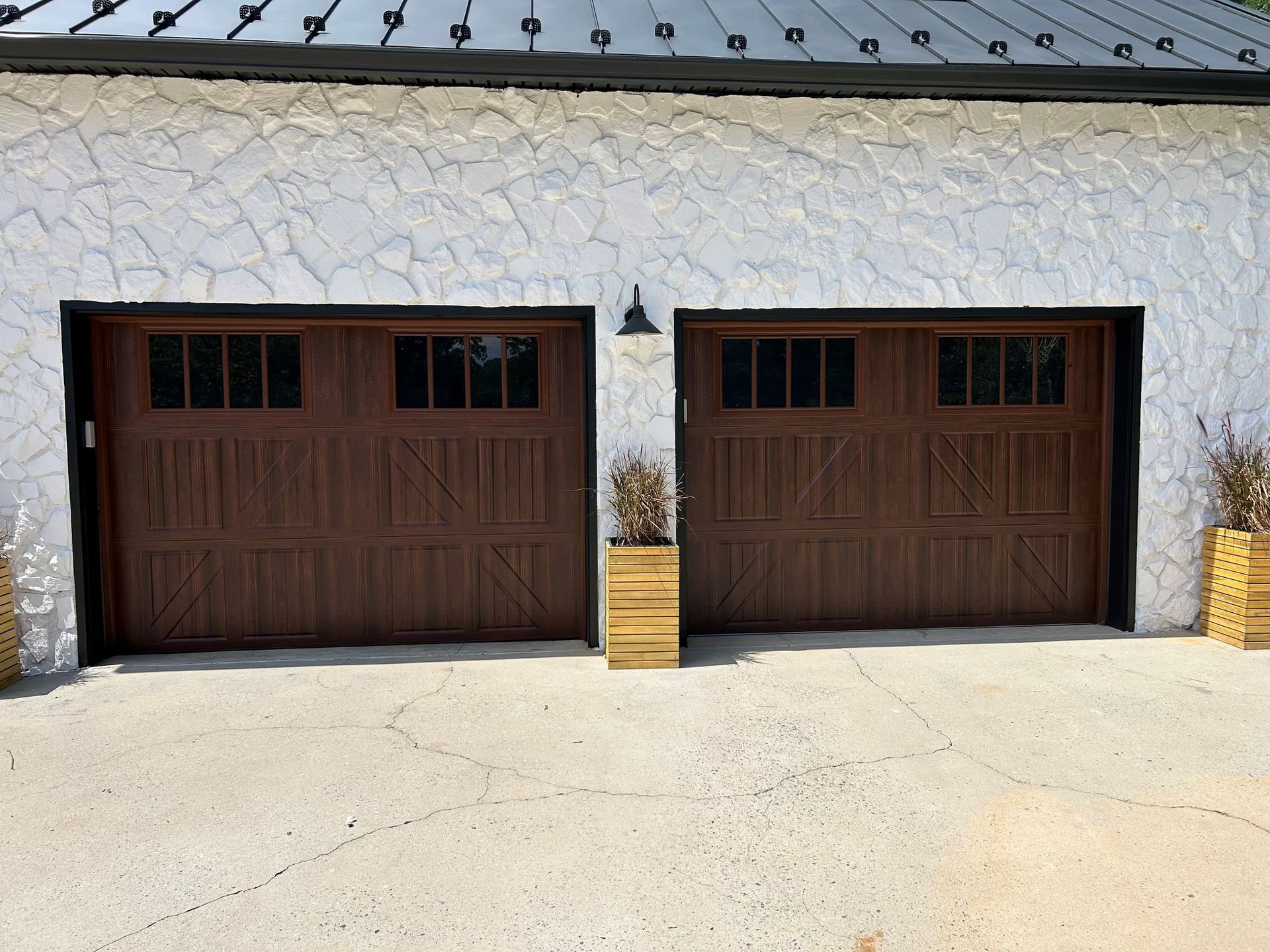 Two brown garage doors on a textured white building. Concrete driveway, lamp, and planters present.