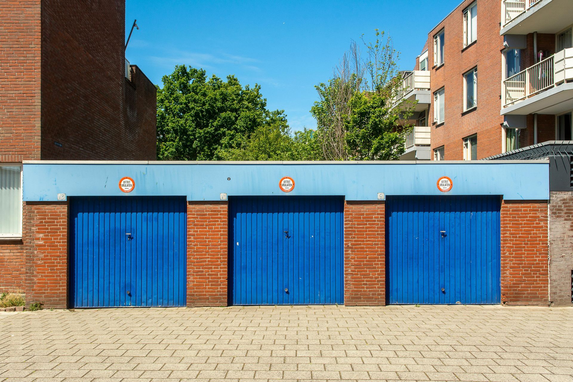 Three blue garage doors with brick accents under a blue stripe against a background of buildings and trees.
