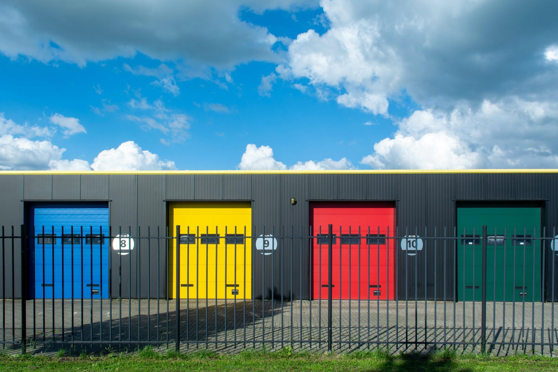 Four storage units with brightly colored doors (blue, yellow, red, green) behind a black fence.