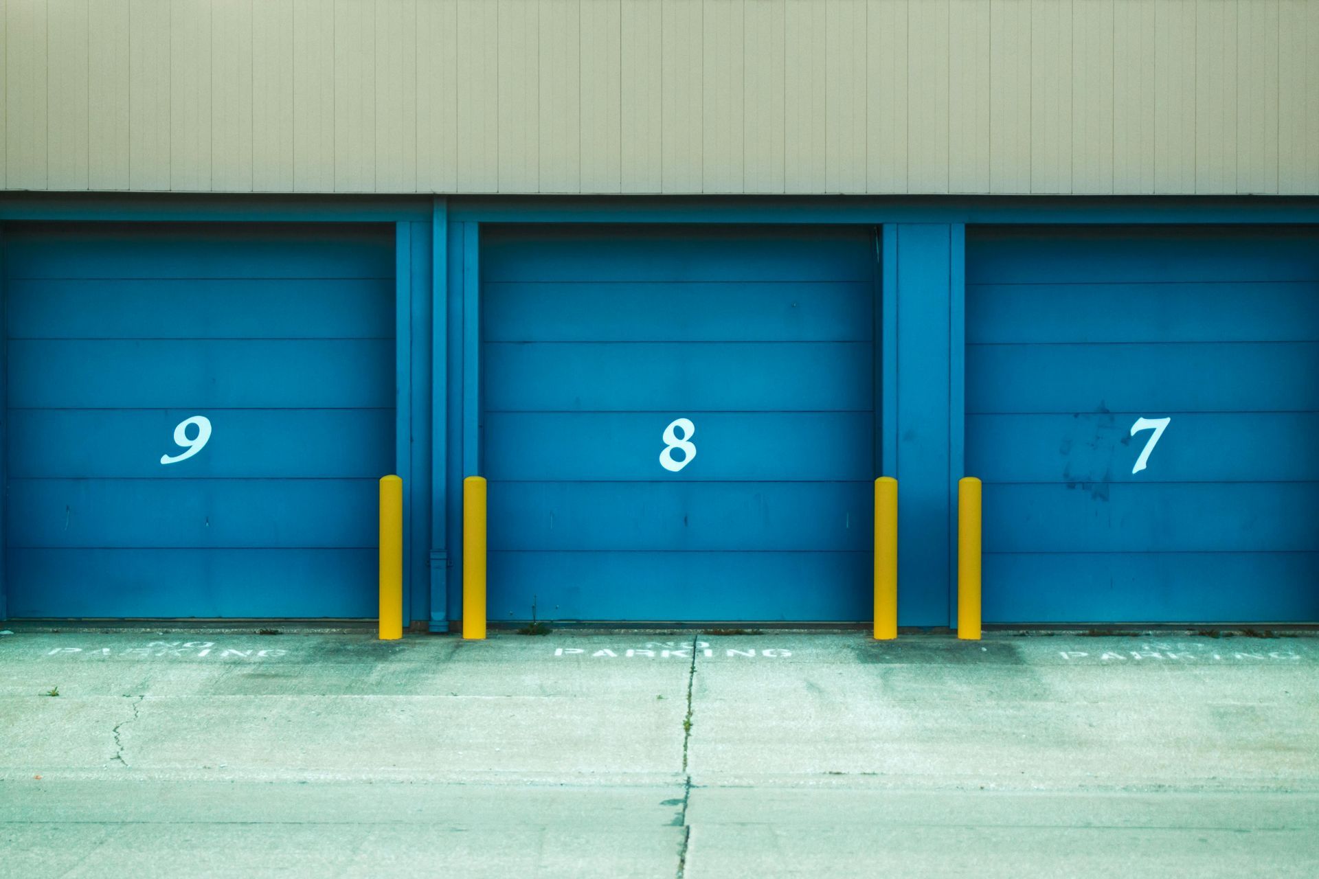 Blue storage unit doors numbered 9, 8, and 7 with yellow bumpers, set in concrete.