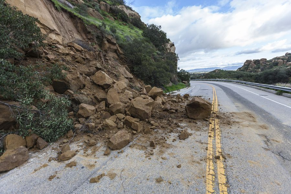 Landslide on a road, blocking a lane. Large rocks and debris cover the asphalt next to a green hillside.