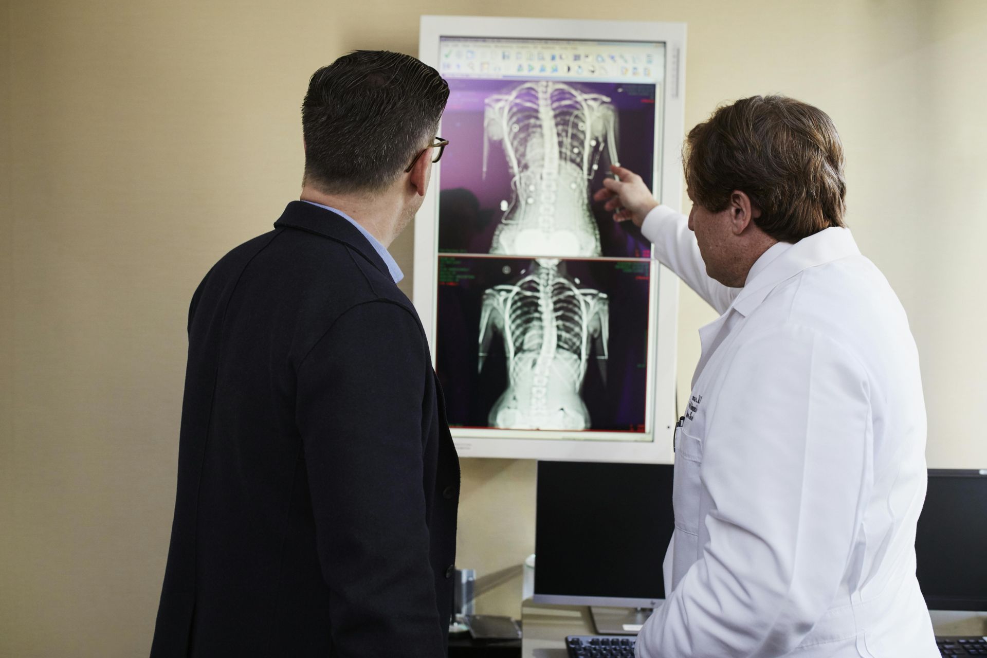 A physician in a white coat points to spinal X-ray images on a light box while discussing them with a person in a suit.