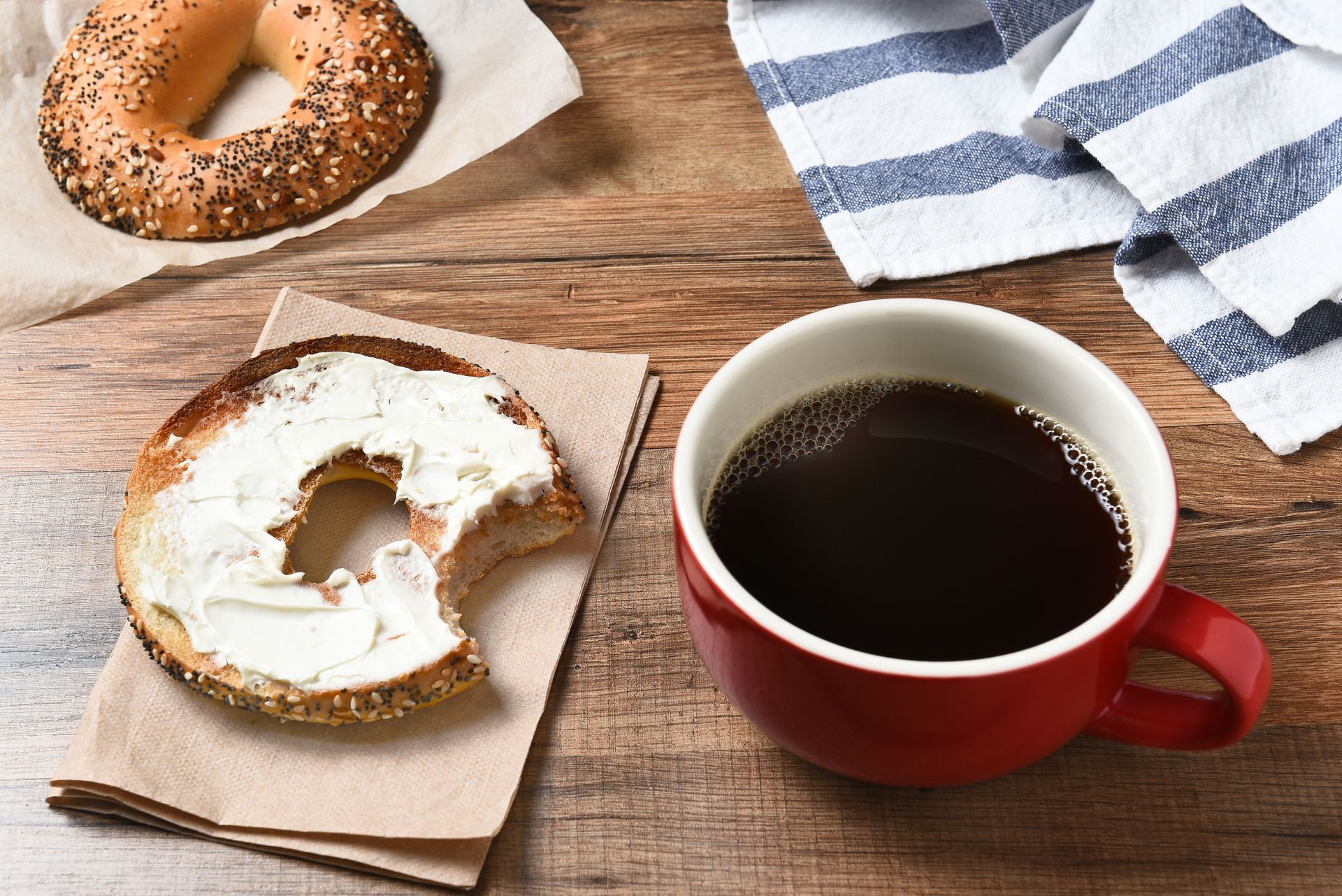 A bagel with cream cheese and a cup of coffee on a wooden table.