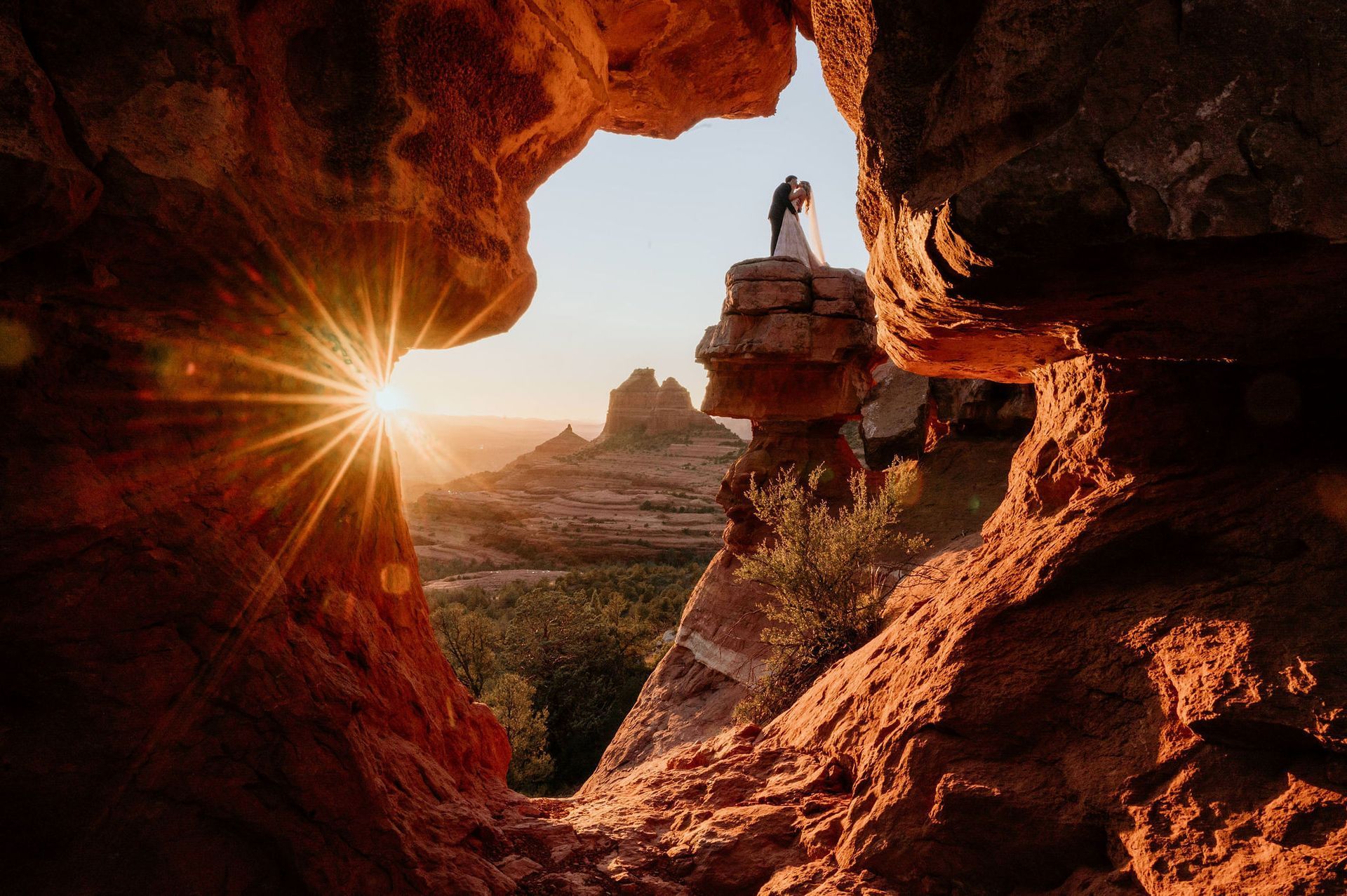 A bride and groom are standing on top of a rocky cliff at sunset.