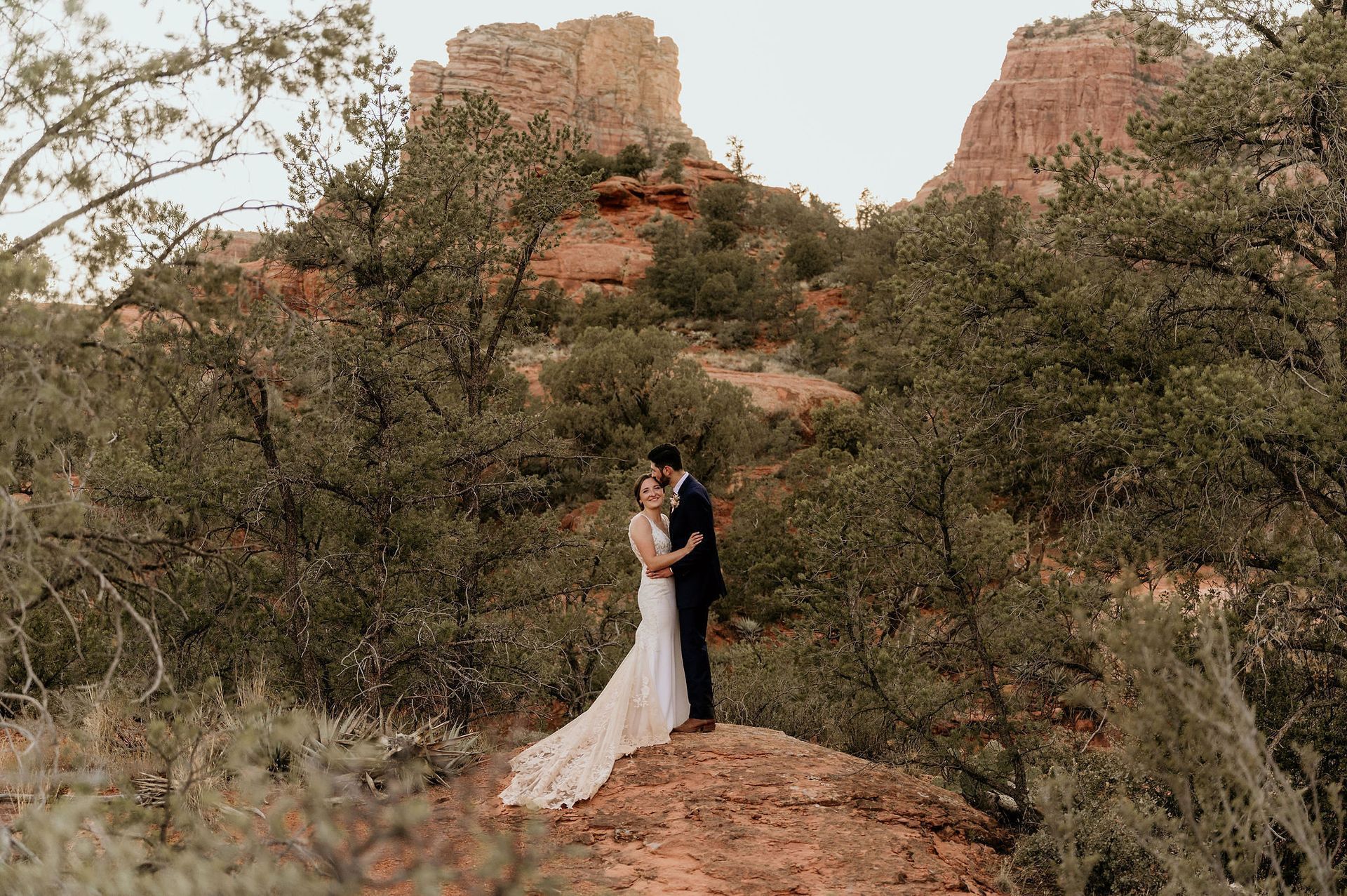 A bride and groom are standing on top of a rock in the woods.
