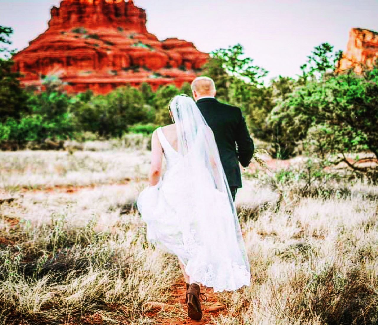 A bride and groom are walking through a field with a mountain in the background.