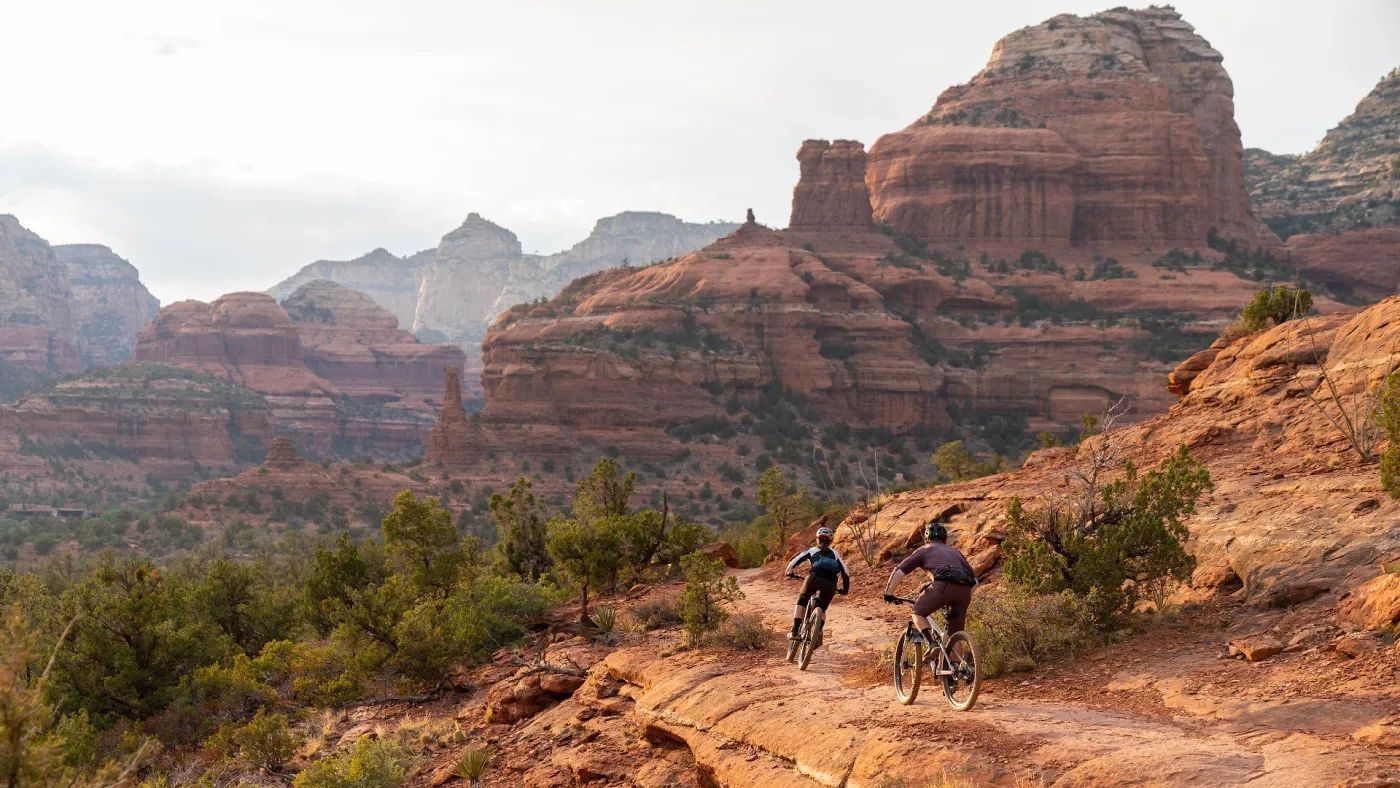 Two people are riding bikes down a dirt trail in the mountains.
