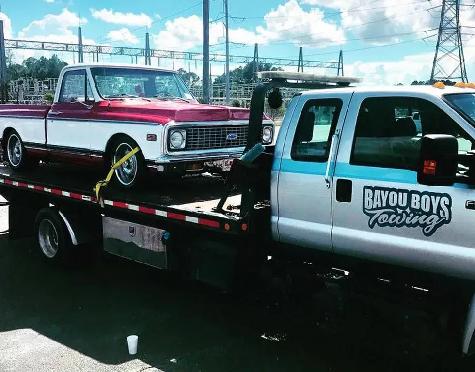 Classic red and white truck being towed by a Bayou Boys towing truck. Outdoors, sunny day.