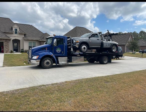 A blue tow truck with two cars on the back is parked in front of a house.