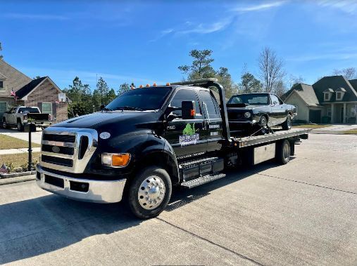 A black tow truck with a car on the back is parked on the side of the road.