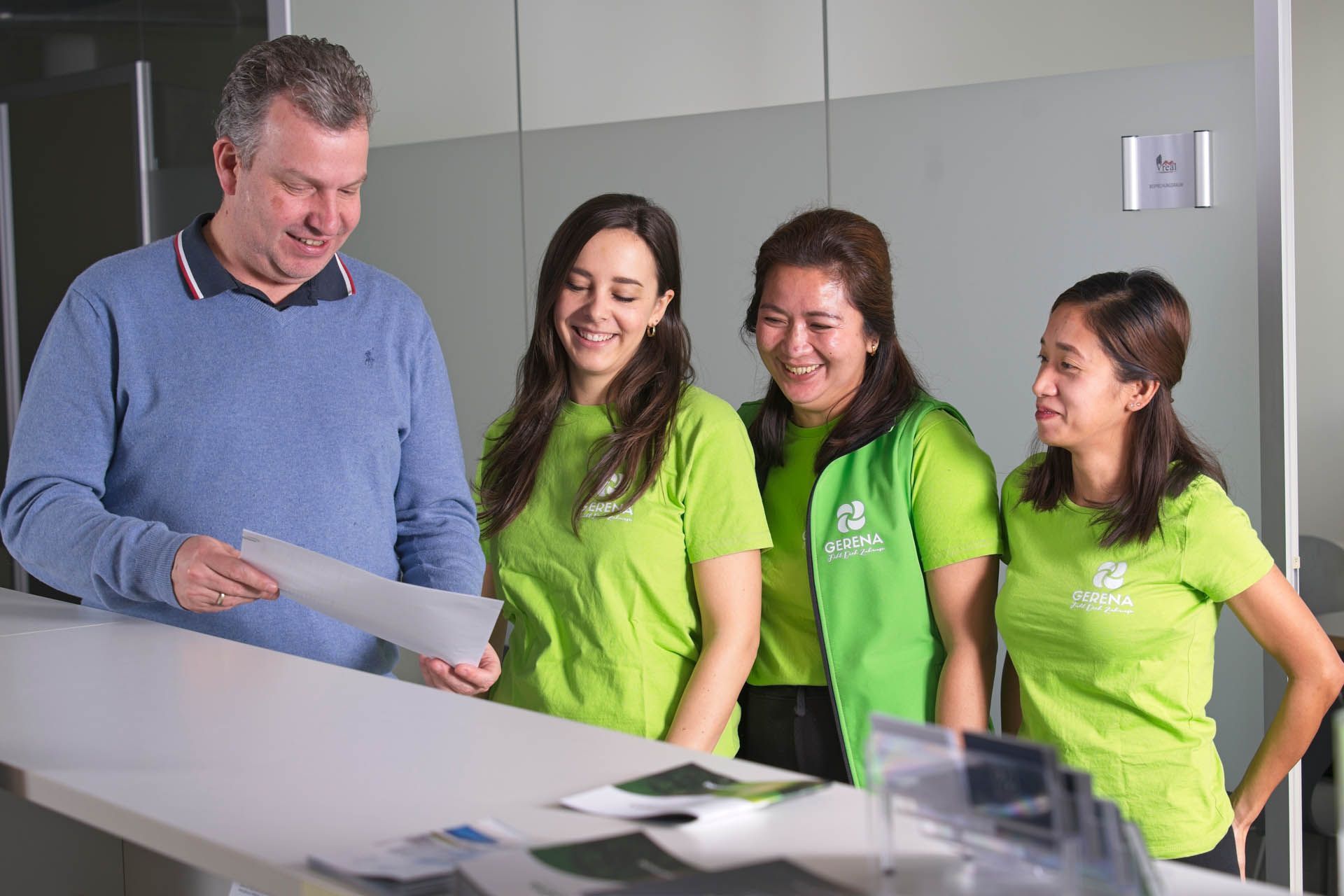 Ein Mann zeigt drei Frauen an einem Schalter ein Dokument; sie lächeln in einer Büroumgebung.