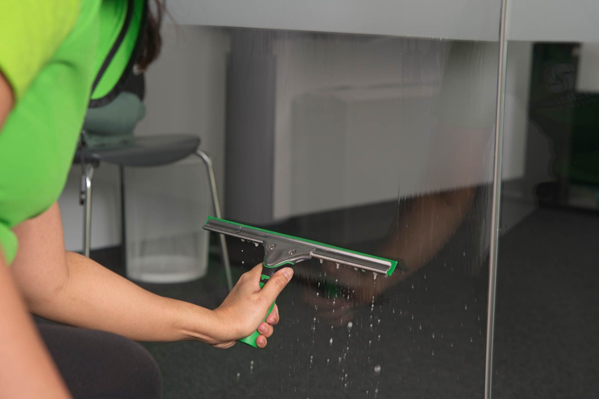 Person wischt eine Glasoberfläche mit einem grünen Abzieher ab; in einem Innenraum.