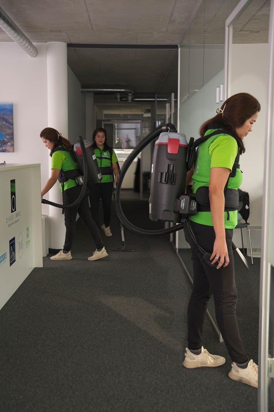 Drei Personen in grünen Hemden und schwarzen Hosen saugen mit Rucksackstaubsaugern ein Büro.