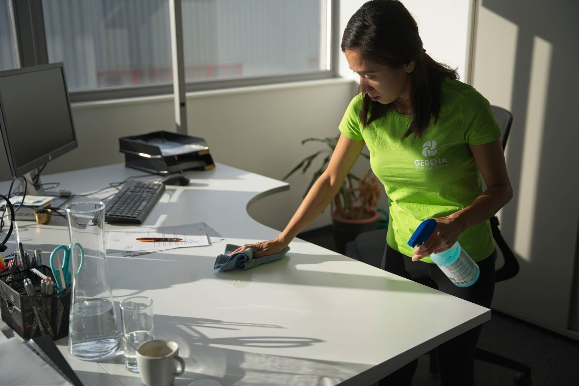 Frau in grünem Hemd reinigt einen weißen Büroschreibtisch mit einer Sprühflasche und einem Tuch.